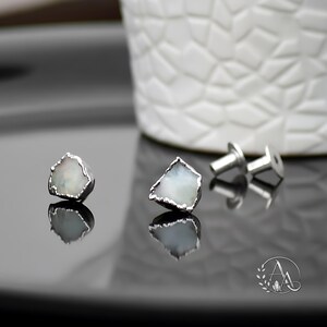 May include: A pair of stud earrings featuring raw, irregular-shaped white stones with iridescent flashes, encased in a textured silver-toned metal setting. The set includes two matching metal earring backs, shown alongside the earrings.