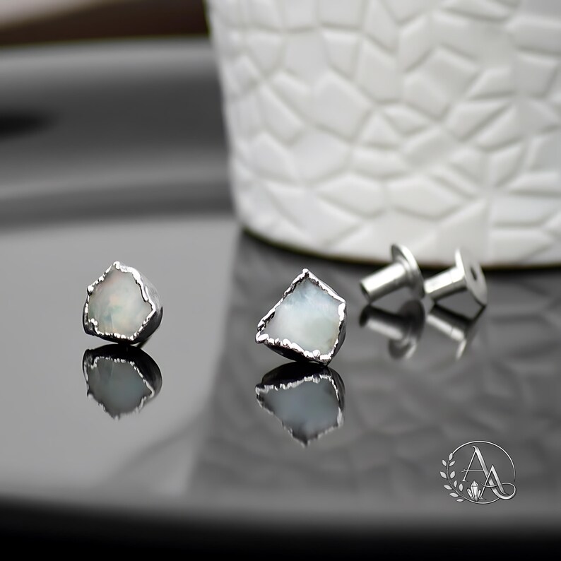 May include: A pair of stud earrings featuring raw, irregular-shaped white stones with iridescent flashes, encased in a textured silver-toned metal setting. The set includes two matching metal earring backs, shown alongside the earrings.