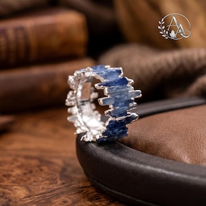 May include: A silver ring featuring a band of raw, jagged blue kyanite crystals set in a textured metal frame. The ring displays a series of vertical, splintered stone segments arranged around the circumference, creating a crown-like appearance.