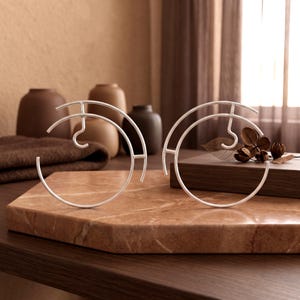 May include: Two silver-toned metal circular stands with an open, abstract design. Each piece features a curved outer frame with a central hook-like element, constructed from thin, smooth wire. The stands are positioned upright on a flat surface.