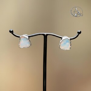 May include: A pair of stud earrings featuring raw, irregular-shaped opals with iridescent flashes of color. Each stone is encased in a thin, textured silver-toned metal setting, showcasing a natural, unpolished aesthetic for these small accessories.