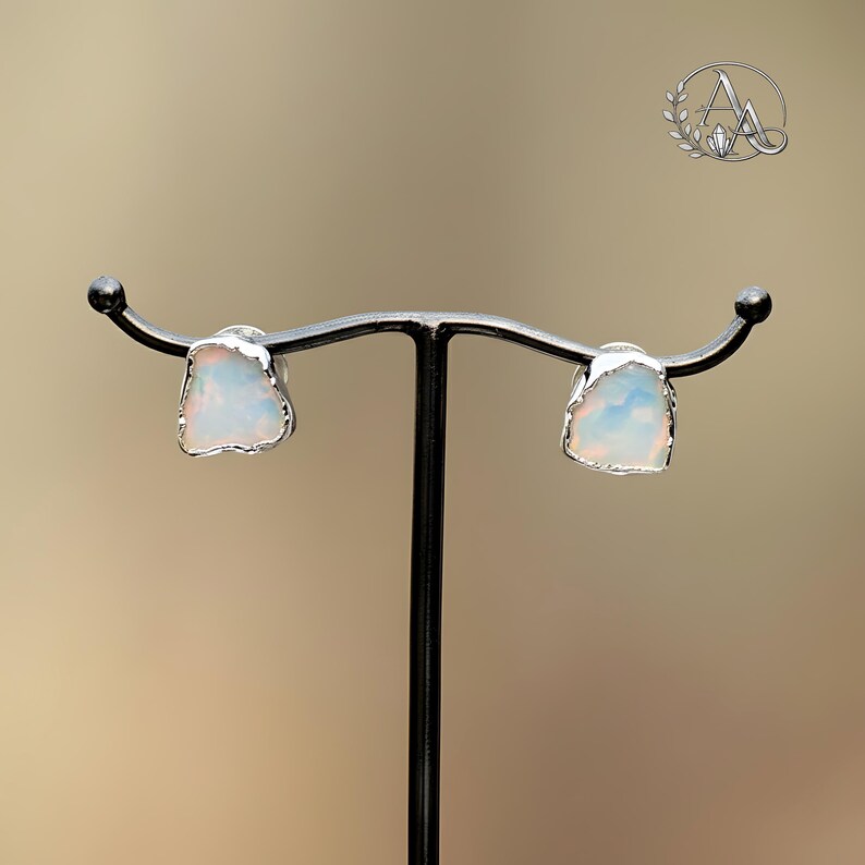 May include: A pair of stud earrings featuring raw, irregular-shaped opals with iridescent flashes of color. Each stone is encased in a thin, textured silver-toned metal setting, showcasing a natural, unpolished aesthetic for these small accessories.