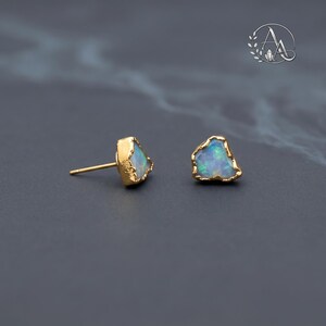 May include: A pair of stud earrings featuring raw, irregular-shaped opals with iridescent blue and green flashes. Each opal is encased in a textured, gold-toned metal setting with a thin post extending from the back of one earring.
