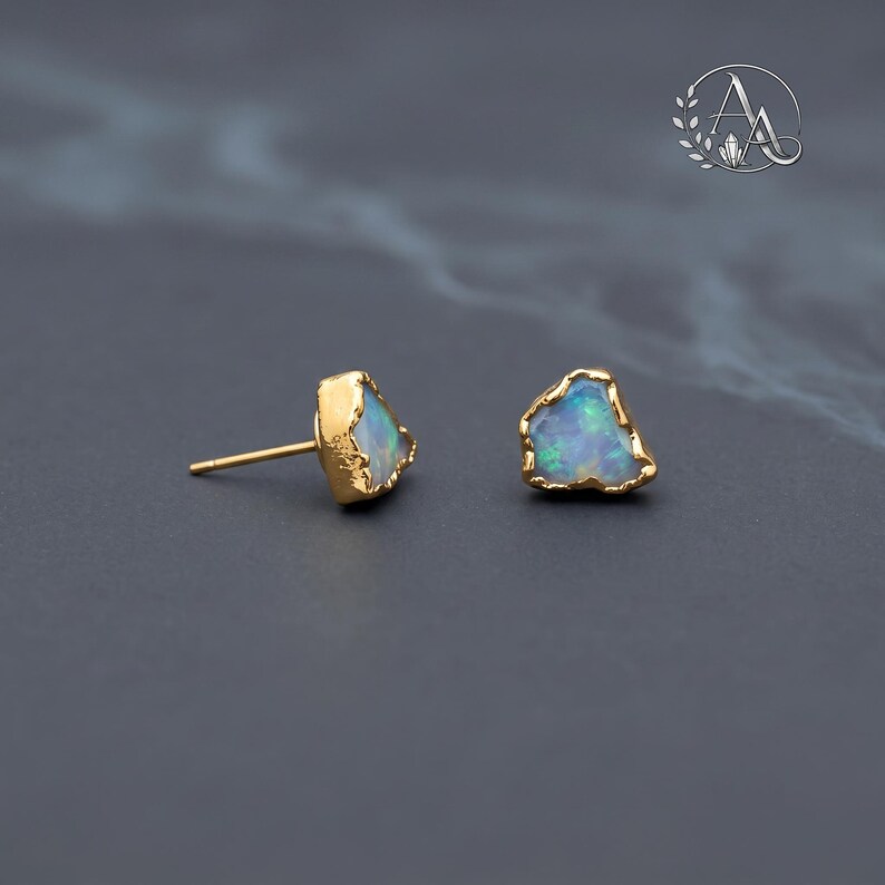 May include: A pair of stud earrings featuring raw, irregular-shaped opals with iridescent blue and green flashes. Each opal is encased in a textured, gold-toned metal setting with a thin post extending from the back of one earring.