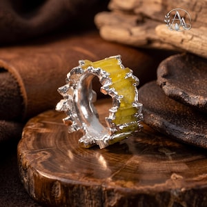 Puede incluir: Un anillo de plata con una banda metálica texturizada y dentada, engastada con una serie de segmentos verticales de piedra amarilla translúcida. El metal tiene un acabado pulido e irregular que enmarca las piedras amarillas.