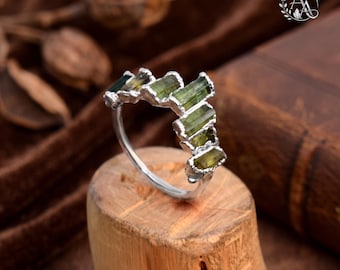 May include: A silver ring featuring a series of raw, rectangular green tourmaline stones set in a staggered, chevron-like arrangement along the top half of the band. The metal band has a polished finish, and the stones display natural variations in olive green hues.