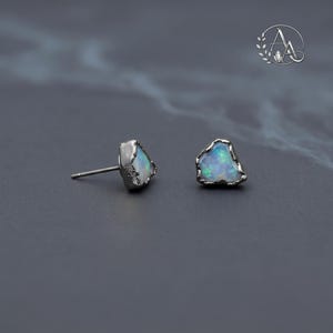 May include: A pair of stud earrings featuring raw, irregular-shaped opals with iridescent blue and green flashes. Each stone is set in a dark, textured metal prong setting. One earring is shown from the side with a post, while the other is front-facing.
