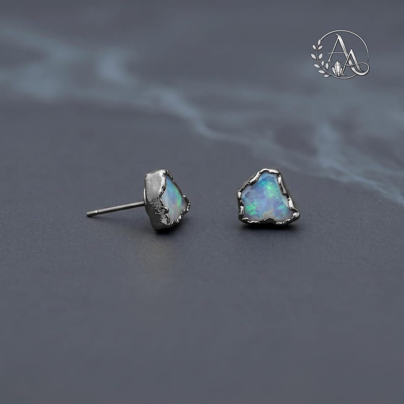 May include: A pair of stud earrings featuring raw, irregular-shaped opals with iridescent blue and green flashes. Each stone is set in a dark, textured metal prong setting. One earring is shown from the side with a post, while the other is front-facing.