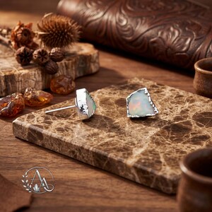 May include: A pair of stud earrings featuring raw, irregular opal gemstones set in silver. Each opal displays a milky white base with iridescent flashes of color. The silver settings have a textured, organic edge that frames the natural shape of the stones.