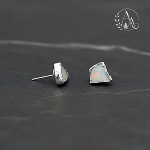 May include: A pair of stud earrings featuring raw, irregular-shaped opals with iridescent flashes of color set in silver-toned metal. One earring is shown from the side displaying the post, while the other shows the front face of the stone.