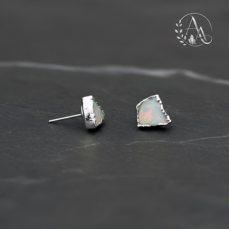 May include: A pair of stud earrings featuring raw, irregular-shaped opals with iridescent flashes of color set in silver-toned metal. One earring is shown from the side displaying the post, while the other shows the front face of the stone.