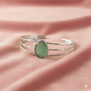 May include: Silver cuff bracelet with a light green oval stone set in the center. The bracelet has three parallel bands and is displayed on a pink fabric surface. A piece of jewelry.