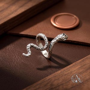 May include: A silver-toned metal ring shaped like a coiled snake with a textured, scale-like surface and a small, dark, circular inlay representing an eye. The design features a wrap-around style with the snake's head and tail extending outward.