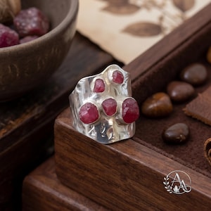 May include: A wide silver ring featuring a hammered, textured surface and four irregular, raw red ruby gemstones set in individual bezels across the front of the band. The metal has a polished finish that reflects light across its uneven surface.