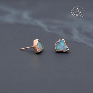 May include: A pair of stud earrings featuring raw, irregular-shaped opals with iridescent blue and green flashes. Each stone is set in a textured rose gold metal bezel with a matching post back, showcasing a natural, unpolished gemstone aesthetic.