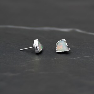 K&ouml;nnte beinhalten: Ein Paar Ohrstecker mit rohen, unregelm&auml;&szlig;ig geformten wei&szlig;en Opalen, die irisierende Farbblitze aufweisen. Jeder Opal ist in eine texturierte, silberfarbene Metallfassung eingefasst, wobei ein d&uuml;nner Stift an der R&uuml;ckseite zur Befestigung dient.