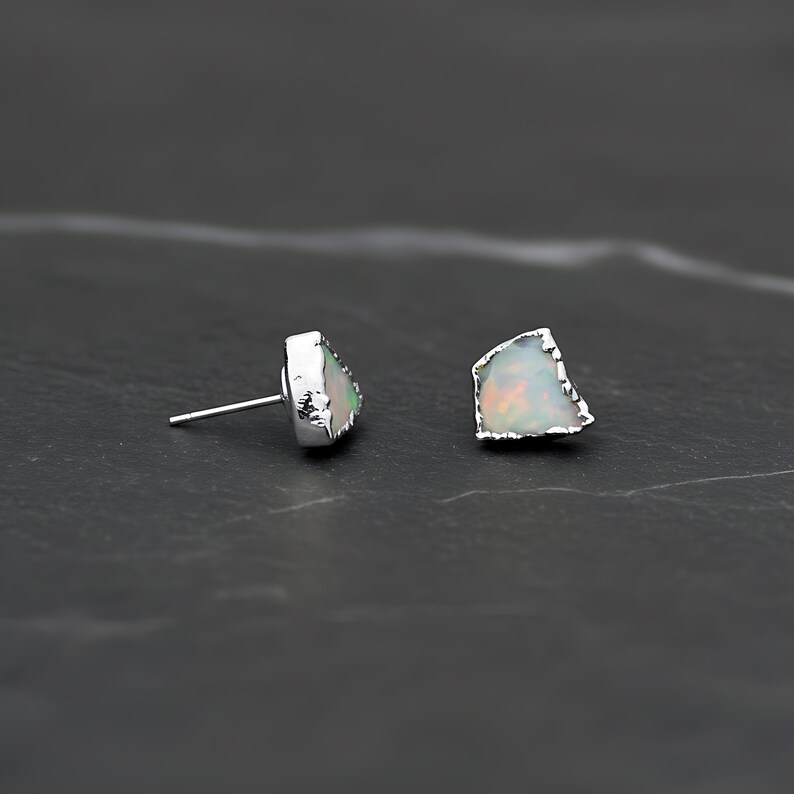 K&ouml;nnte beinhalten: Ein Paar Ohrstecker mit rohen, unregelm&auml;&szlig;ig geformten wei&szlig;en Opalen, die irisierende Farbblitze aufweisen. Jeder Opal ist in eine texturierte, silberfarbene Metallfassung eingefasst, wobei ein d&uuml;nner Stift an der R&uuml;ckseite zur Befestigung dient.