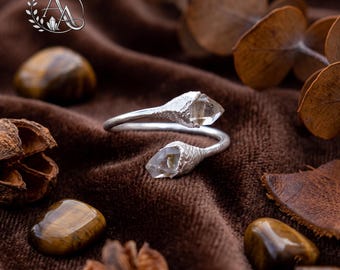 Op de afbeelding: Een zilveren bypass-ring met twee ruwe, heldere kwartspunten gezet in getextureerde, organisch ogende metalen kapjes. De ringband heeft een gladde, gepolijste afwerking en het ontwerp is verstelbaar, waarbij de twee kristalpunten naar elkaar toe wijzen.
