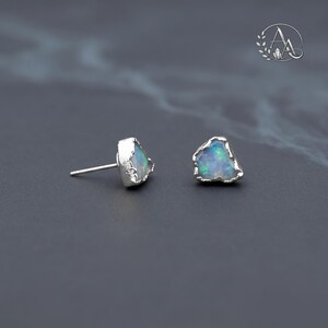 May include: A pair of stud earrings featuring raw, irregular opal stones with iridescent blue and green flashes, set in textured silver metal settings. Each earring has a silver post back. The stones have a natural, unpolished appearance.