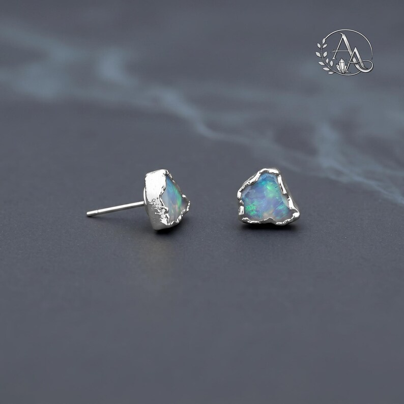 May include: A pair of stud earrings featuring raw, irregular opal stones with iridescent blue and green flashes, set in textured silver metal settings. Each earring has a silver post back. The stones have a natural, unpolished appearance.