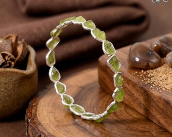Puede incluir: Un brazalete circular que presenta una serie de pequeñas piedras de peridoto en bruto de color verde claro, engastadas en un fino marco de metal plateado. Las piedras están encajadas individualmente y unidas formando un aro continuo.