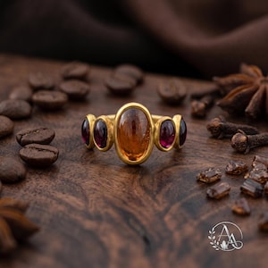 May include: A gold ring featuring a large, central oval-shaped amber-colored gemstone flanked by two smaller, deep red oval-shaped gemstones on each side. The metal band has a smooth, matte finish, and the stones are set in simple gold bezels.