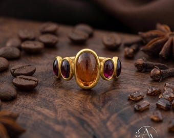 May include: A gold ring featuring a large, central oval-shaped amber-colored gemstone flanked by two smaller, deep red oval-shaped gemstones on each side. The metal band has a smooth, matte finish, and the stones are set in simple gold bezels.