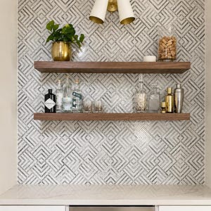 Pode incluir: Duas prateleiras horizontais de madeira montadas contra uma parede com um padr&atilde;o geom&eacute;trico de azulejos cinzentos e brancos. A prateleira superior cont&eacute;m um vaso dourado com uma planta verde e um frasco de vidro cheio de rolhas.
