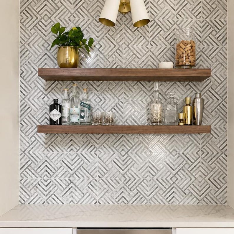 Pode incluir: Duas prateleiras horizontais de madeira montadas contra uma parede com um padr&atilde;o geom&eacute;trico de azulejos cinzentos e brancos. A prateleira superior cont&eacute;m um vaso dourado com uma planta verde e um frasco de vidro cheio de rolhas.