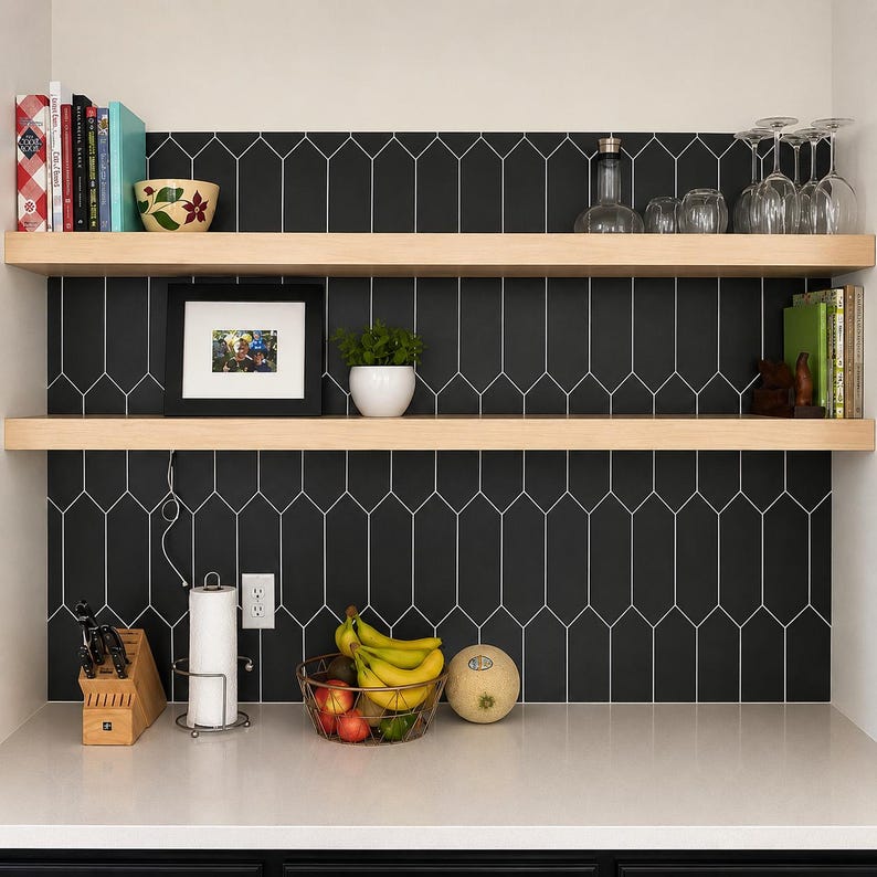 Pode incluir: Duas prateleiras flutuantes de madeira clara montadas contra um revestimento de azulejos pretos em formato de estaca. As prateleiras cont&ecirc;m livros, uma tigela floral, copos, uma foto emoldurada, uma pequena planta e um bloco de madeira para facas.