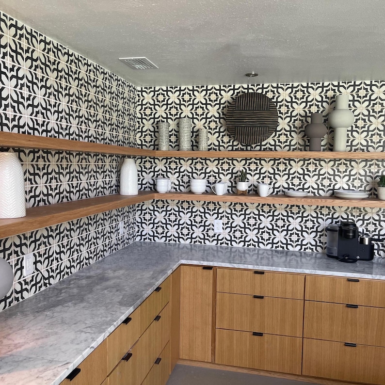 Pode incluir: Uma cozinha com arm&aacute;rios de madeira clara com ferragens pretas, bancada de m&aacute;rmore cinza e prateleiras de madeira abertas. As paredes s&atilde;o revestidas com azulejos quadrados com padr&otilde;es em preto e branco. V&aacute;rios vasos e canecas est&atilde;o expostos.