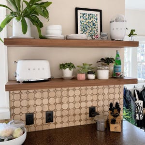 Pode incluir: Duas prateleiras horizontais de madeira montadas numa parede com um revestimento de azulejos geom&eacute;tricos bege. As prateleiras cont&ecirc;m uma torradeira Smeg branca, um espremedor de citrinos Smeg branco, v&aacute;rias plantas verdes e pratos de cer&acirc;mica.
