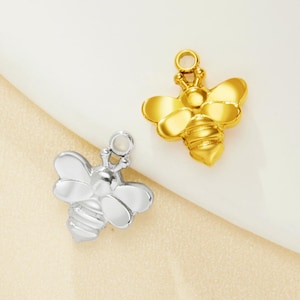 May include: Two bee-shaped charms, one silver and one gold, are displayed against a soft, off-white background. The charms have detailed wings and bodies, with a small loop at the top for attachment. The gold charm has a shiny finish, while the silver charm has a polished look.