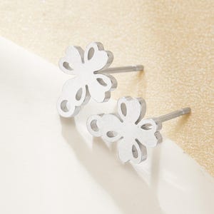 May include: A pair of silver-colored stud earrings. Each earring features a clover-shaped design with four rounded petals and a post for insertion. The earrings are displayed on a white surface with a light beige background.
