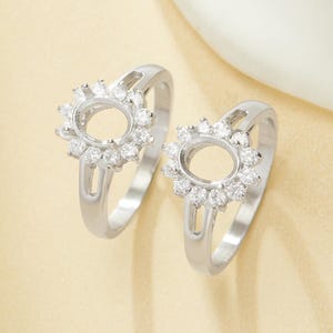 Puede incluir: Dos anillos de plata con diseño floral. Cada anillo presenta un engaste central ovalado rodeado de pequeñas piedras brillantes. Las bandas son lisas y sencillas, con un diseño dividido cerca del engaste. Los anillos se exhiben sobre una superficie amarillo claro.