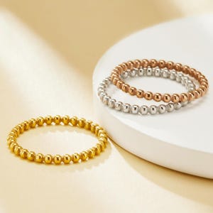 Puede incluir: Tres anillos de cuentas en oro, plata y oro rosa. Los anillos están apilados sobre una superficie blanca y dispuestos sobre un fondo amarillo claro. Los anillos están hechos de pequeñas cuentas redondas.