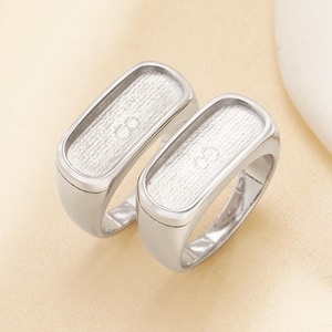 May include: Two silver-toned rings with rectangular settings. The settings have a textured surface and a curved, rounded band. The rings are displayed on a light beige surface. The rings are likely for jewelry making or crafting.