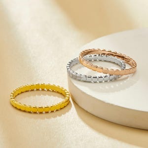 May include: Three stackable rings in gold, silver, and rose gold. Each ring features a repeating square design. The rings are displayed on a white surface with a soft, neutral background.