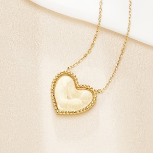 Op de afbeelding: Een goudkleurige ketting met een hartvormige hanger. De hanger heeft een crèmekleurig midden en een kralenrand. De fijne ketting is ook goudkleurig. De achtergrond is zacht off-white, wat een eenvoudige, elegante uitstraling creëert.