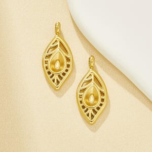 May include: Two gold-colored pendant earrings. Each earring has a teardrop-shaped design with an openwork leaf pattern and a central teardrop-shaped element. The earrings are displayed on a light-colored surface.