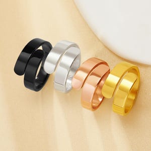May include: Four spiral rings in black, silver, rose gold, and gold. Each ring has a wrap-around design, creating a modern look. The rings are displayed on a light beige surface, showcasing their metallic finishes.
