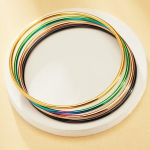 Puede incluir: Una colección de brazaletes finos y coloridos en tonos dorados, verdes, cobrizos, negros e iridiscentes. Las pulseras están dispuestas en un patrón circular sobre una superficie blanca y redonda, creando una exhibición vibrante de accesorios.