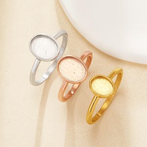 May include: Three rings with oval settings in silver, rose gold, and gold. The rings have a simple band design and are set against a neutral background. The settings are designed to hold a stone or other decorative element.