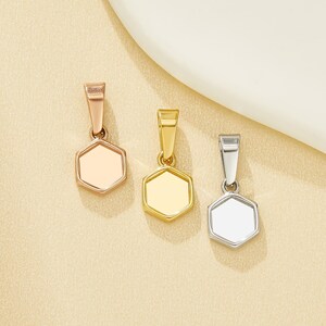 Può includere: Tre ciondoli esagonali in oro rosa, oro e argento. Ogni ciondolo ha un piccolo anello nella parte superiore per una catena. I ciondoli sono esposti su una superficie beige chiaro.
