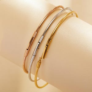 Pode incluir: Três pulseiras de metal em ouro rosa, prata e ouro. Cada pulseira tem um design simples e curvo com fecho. As pulseiras são exibidas em um fundo neutro, mostrando seu estilo elegante e minimalista.