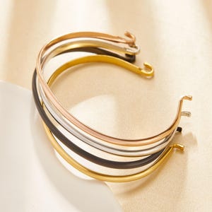 Puede incluir: Una colección de seis pulseras de metal en oro, plata, oro rosa y negro. Las pulseras tienen un diseño simple y curvado con un cierre de gancho. Las pulseras están apiladas, mostrando sus diferentes acabados.