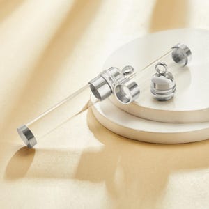 Peut inclure: Un flacon en verre transparent avec des accents métalliques argentés, comprenant un bouchon à vis et des anneaux décoratifs. Le flacon est cylindrique et semble vide. Les composants métalliques ont une finition polie. Le flacon est présenté sur une plateforme blanche et circulaire.