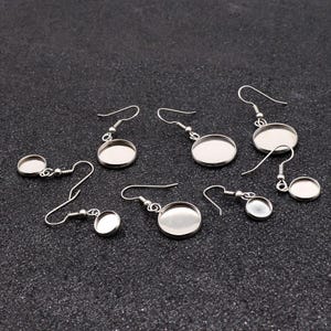 May include: A set of six silver earring blanks with round, flat, silver-toned metal settings. The settings are designed to hold a variety of materials, such as resin, glass, or fabric.