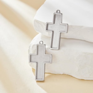 May include: Two silver metal cross charms with a hollow center. The charms are on a white background.