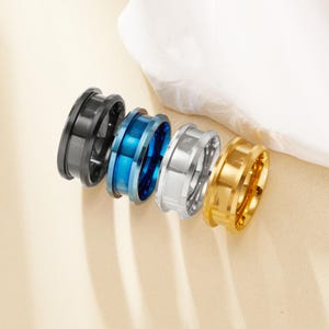 May include: Four rings in a row, each with a unique color. The rings are black, blue, silver, and gold. The rings have a unique design with a recessed center and a raised outer edge. The rings are made of metal and have a polished finish.